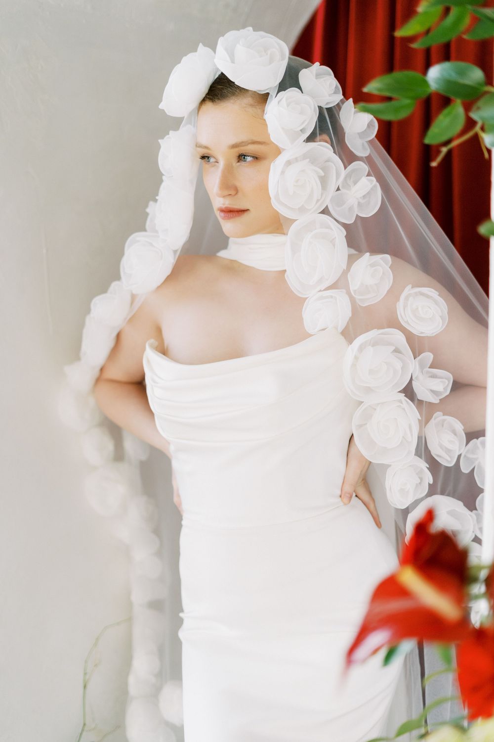 Bridal portrait with white rose-adorned veil and elegant strapless dress against red curtain.