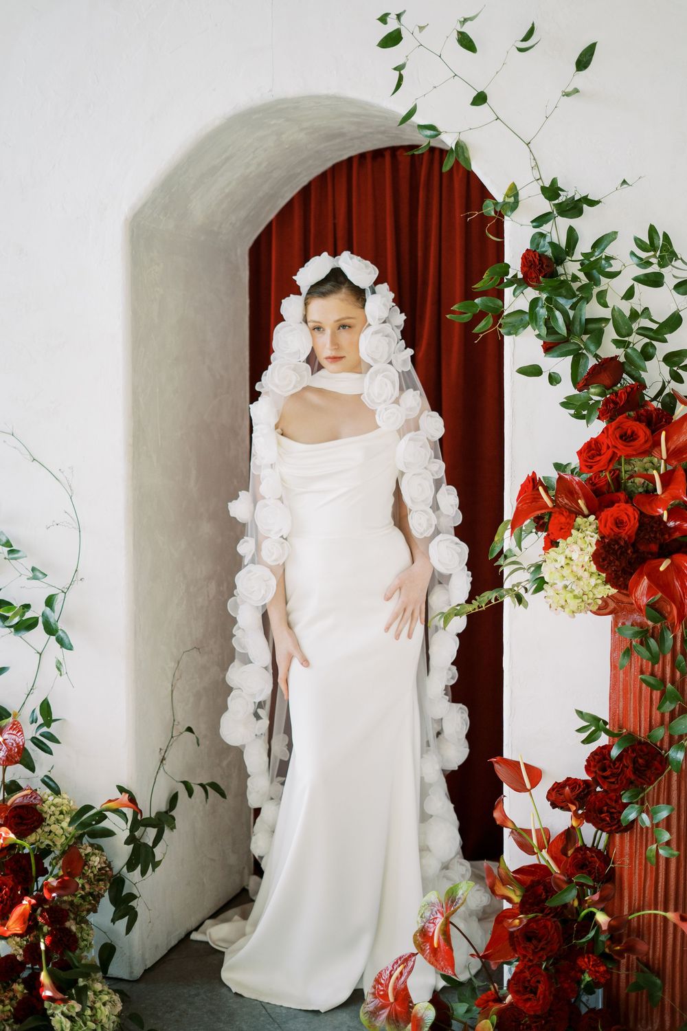 Bride poses in fitted white gown with unique pom-pom adorned veil against arch with red floral arrangements.