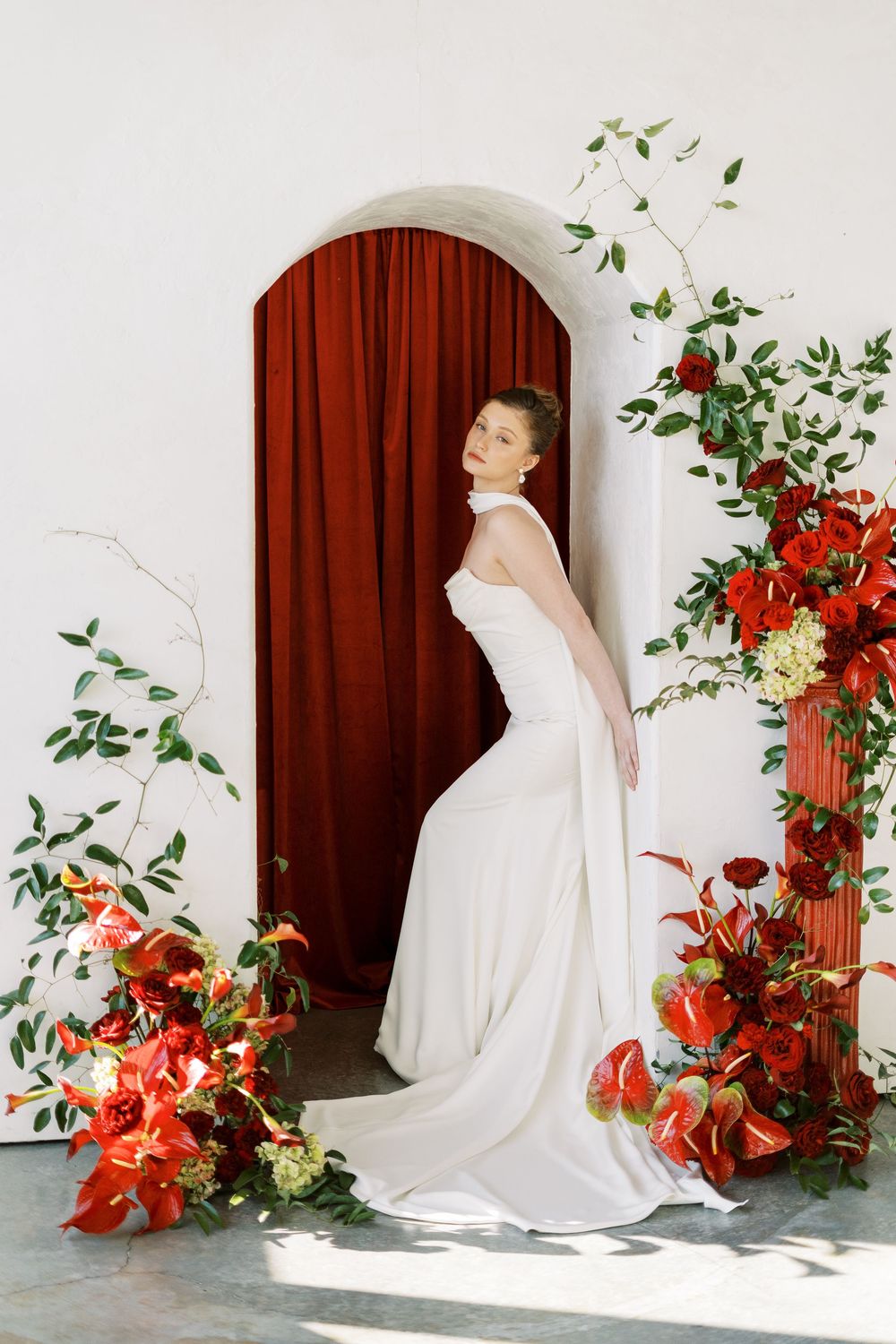 Elegant bridal scene with white gown against arched doorway surrounded by red florals and greenery.