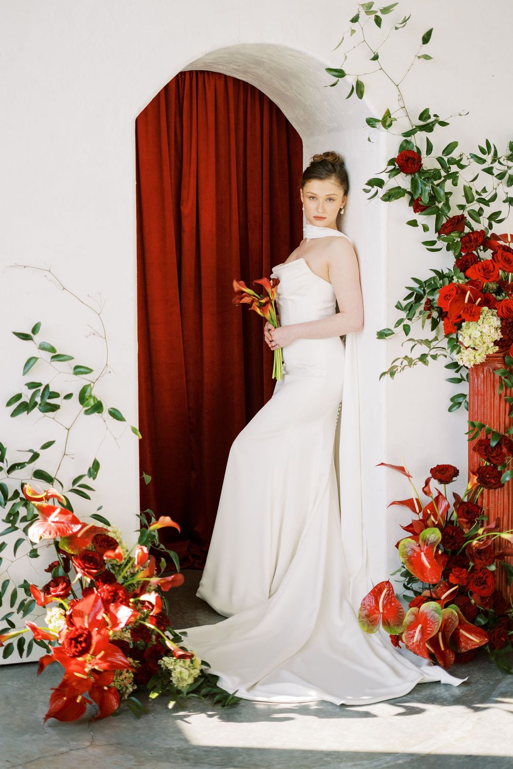 Bridal photography series with white gown against burgundy backdrop with red lilies and green vine decorations.