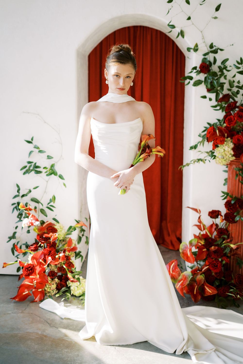 Modern bridal gown with fitted silhouette against red curtain and floral arch backdrop.