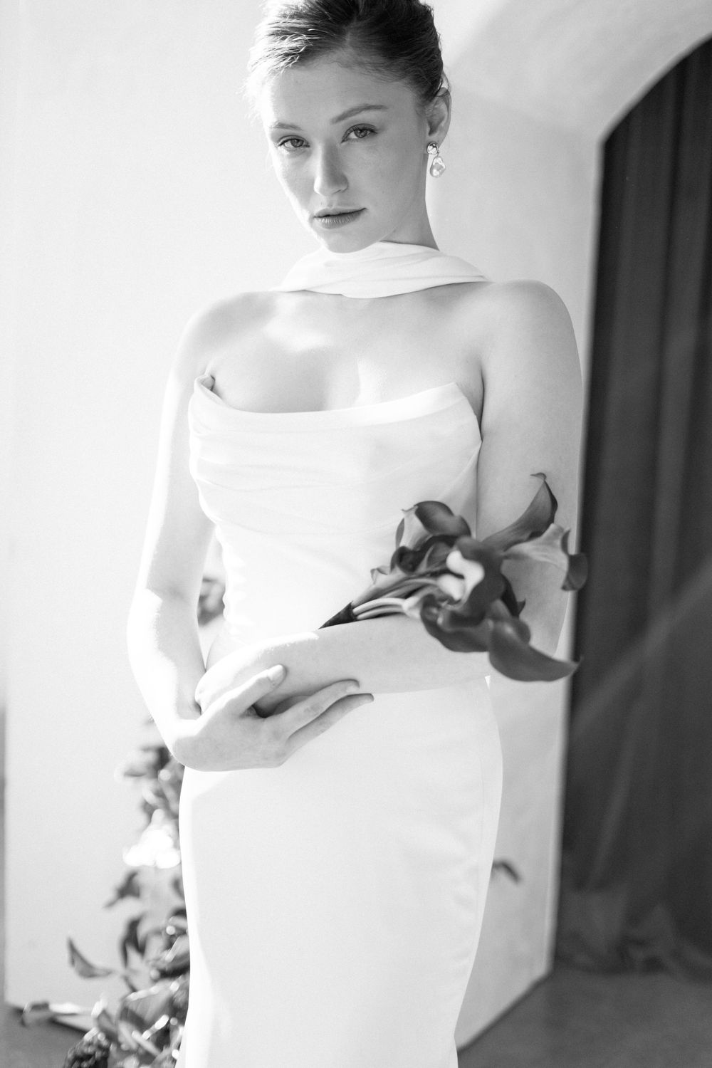 A black and white photo of someone posing in an elegant strapless white wedding dress while holding flowers.
