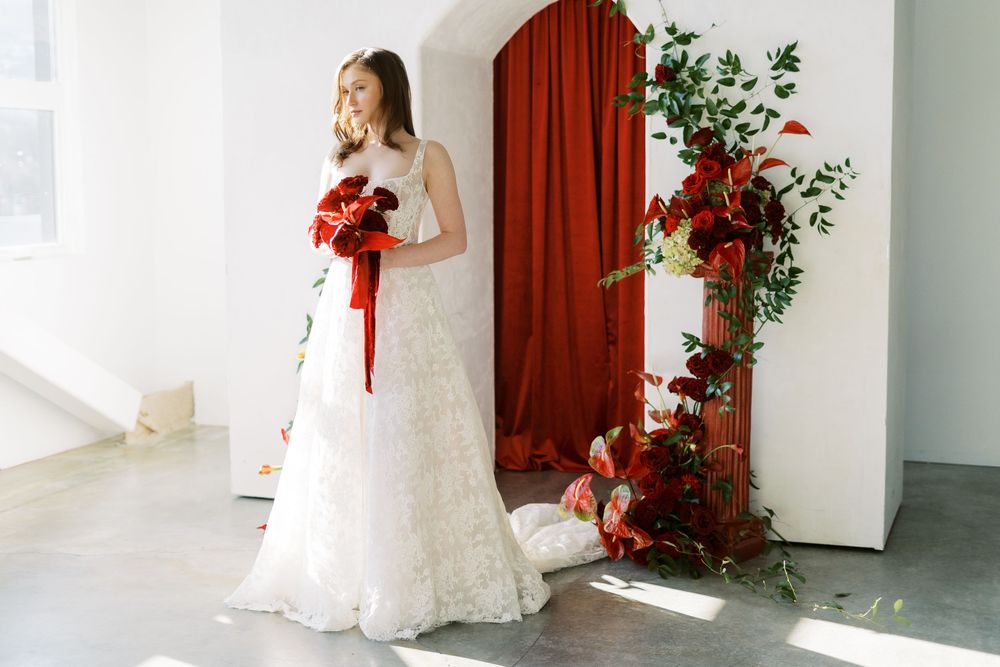 A person in a white lace wedding dress stands against a red curtain backdrop with natural lighting from windows.