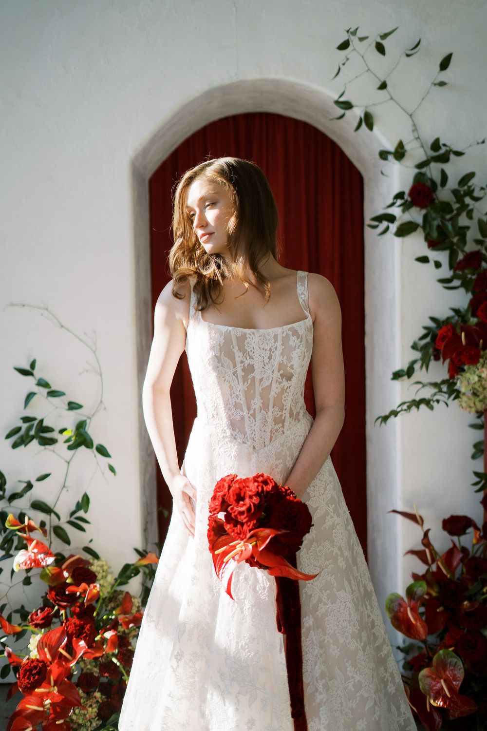 A figure in a white lace wedding dress poses against a wall with red flowers and green vines.