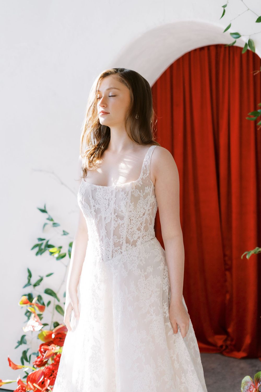 Elegant white lace wedding dress with corset bodice against red curtain backdrop and floral arrangements.