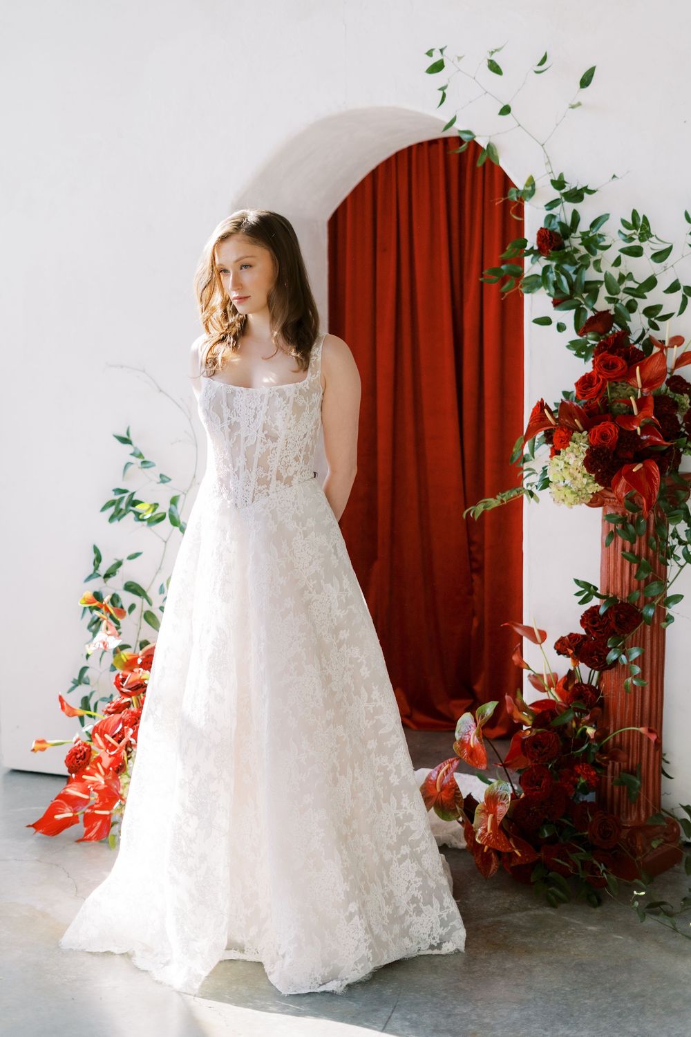 Elegant white lace wedding gown with corset bodice shown against white wall with red curtain and floral decorations.