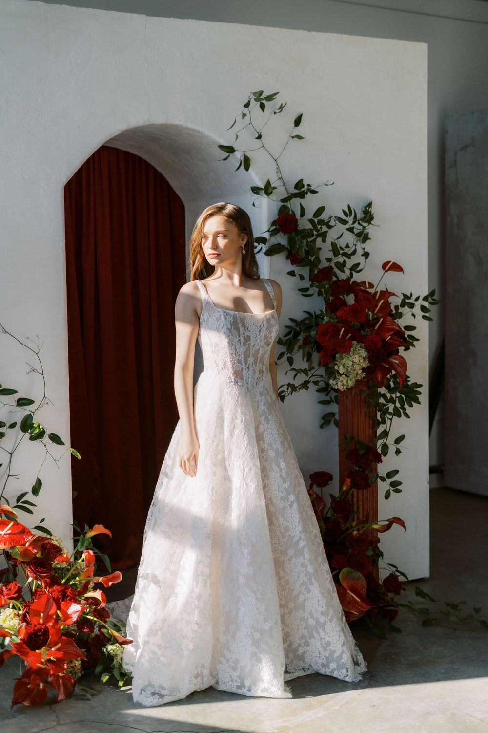 An elegant white lace wedding gown with off-shoulder design photographed against an archway with red floral decorations.