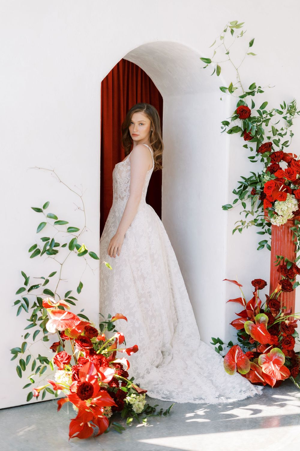 Bride in flowing lace gown poses in archway decorated with red and coral floral arrangements.