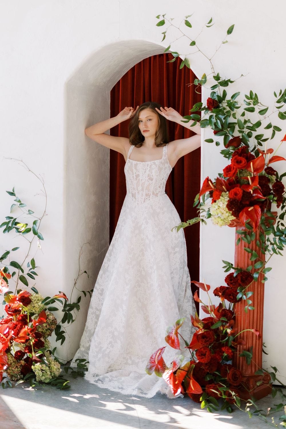 Elegant white lace wedding gown with dramatic floral arch and red flower arrangements against a white wall.