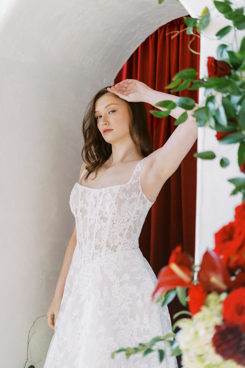 Elegant white lace wedding dress styled with delicate floral details and red curtain backdrop with greenery.