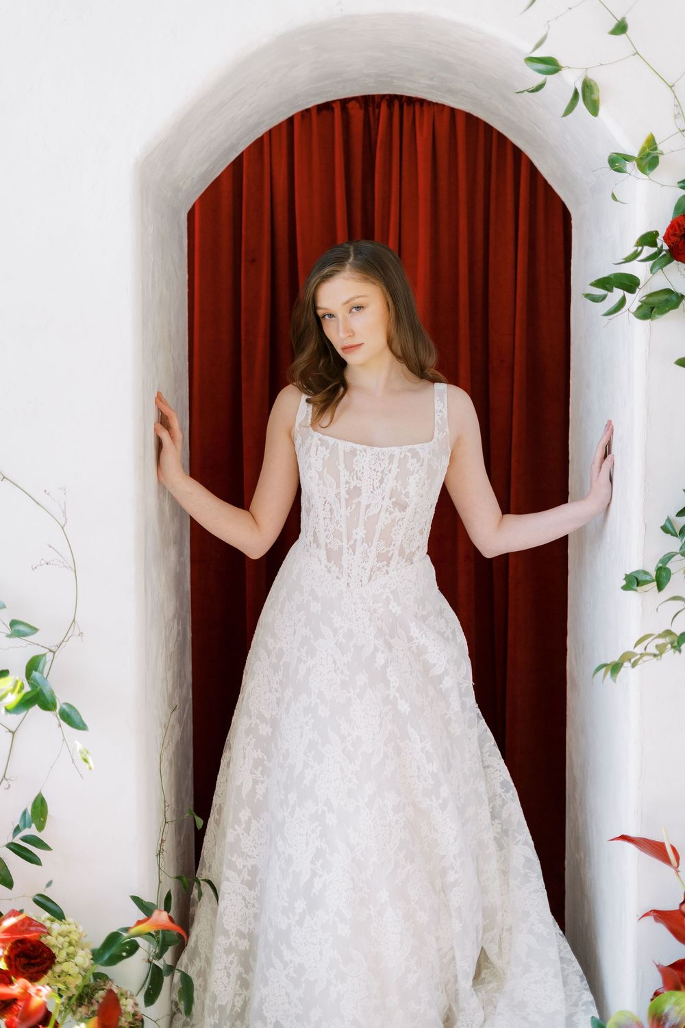 Romantic bridal portrait showcasing a detailed white lace wedding gown framed by a red curtained archway.