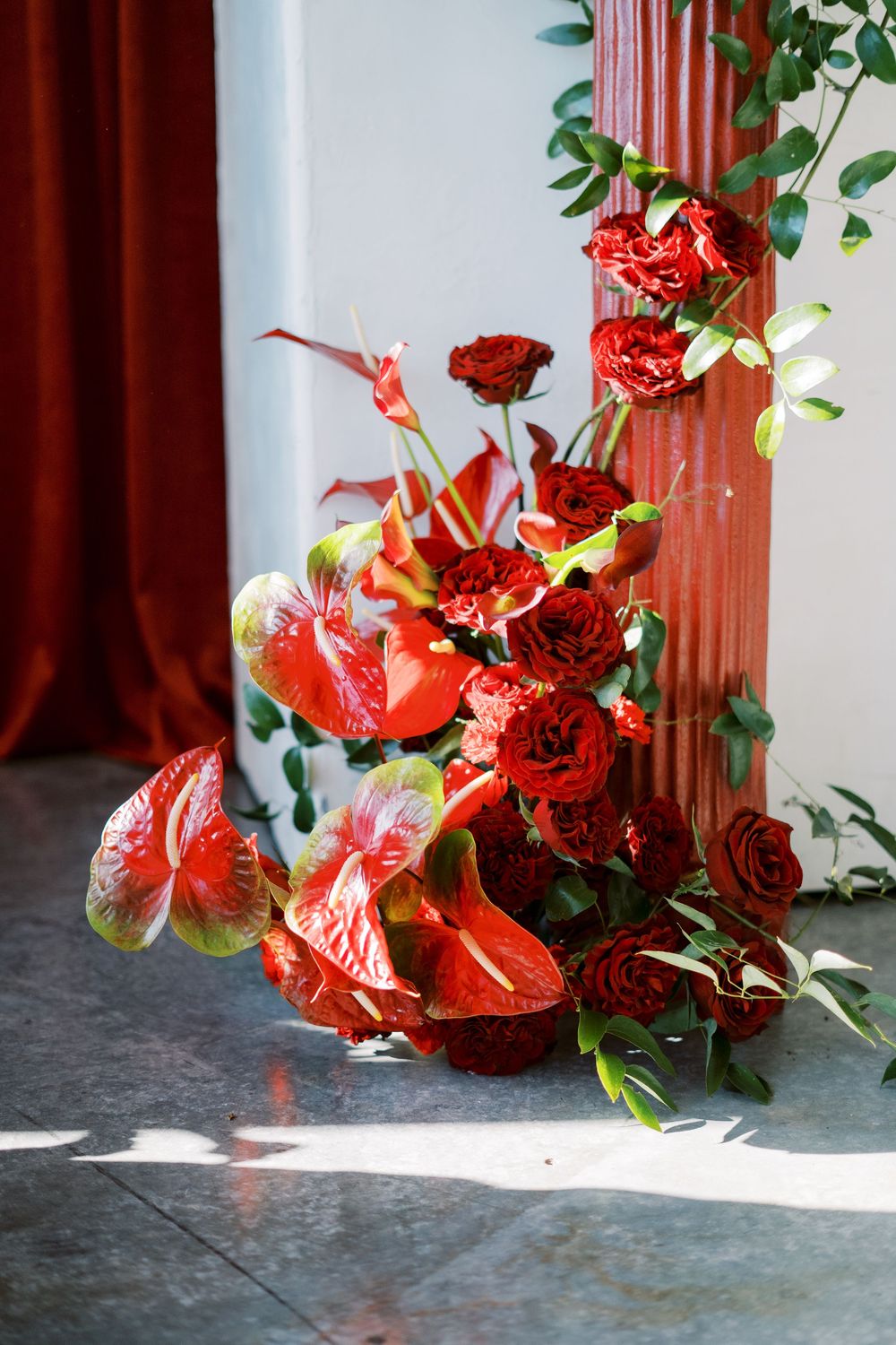 A vibrant floral arrangement with red anthurium lilies and roses sits elegantly against a white wall and wooden column.