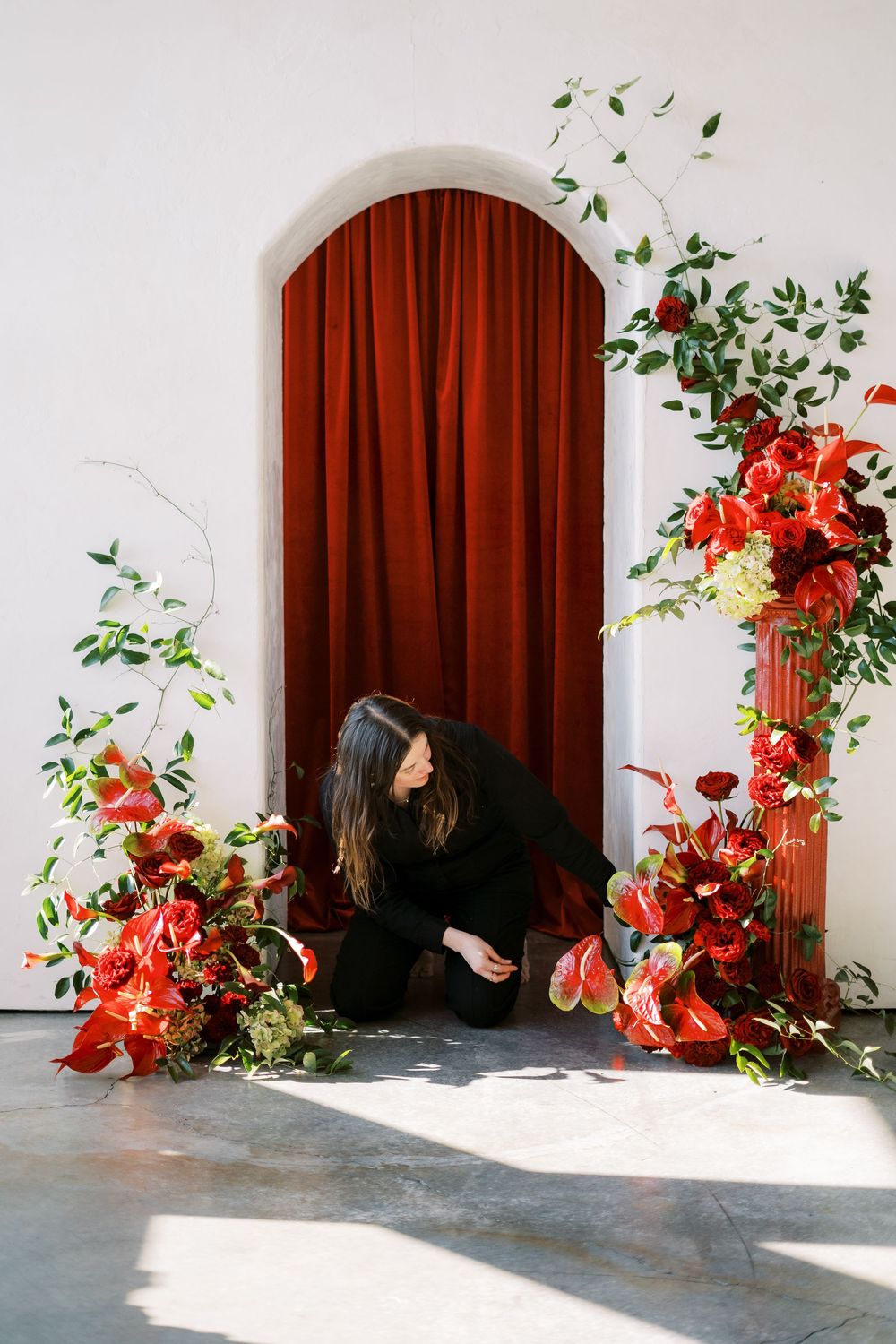 A person in black arranges vibrant red floral decorations around an arched doorway with a burgundy velvet curtain.