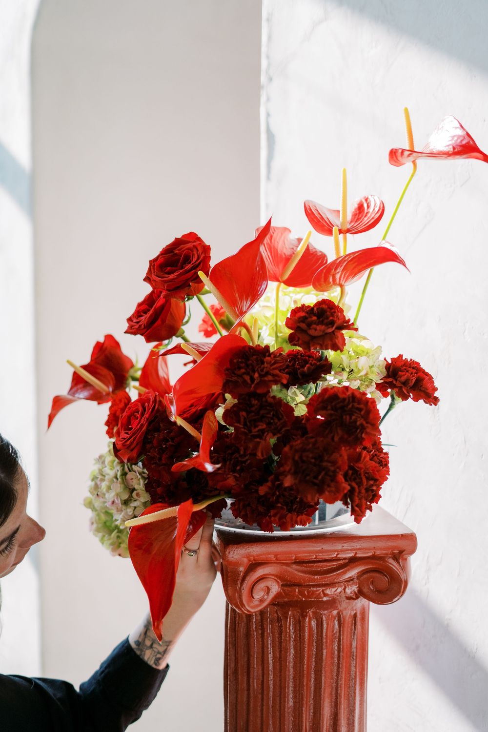 Red roses and anthurium flowers arranged on a decorative terracotta column pedestal against a white wall.