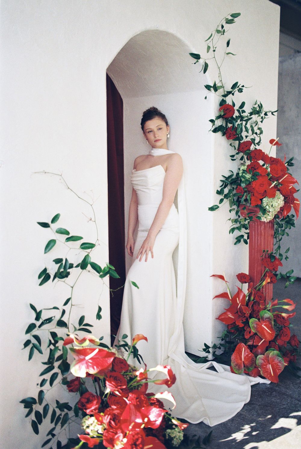 A white strapless gown stands elegantly in an archway decorated with cascading red flowers and green foliage.