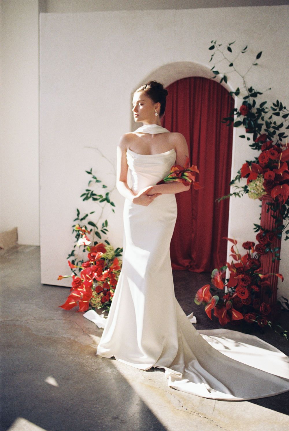 A figure in a flowing white wedding gown twirls elegantly in a sunlit room decorated with vibrant red floral arrangements.