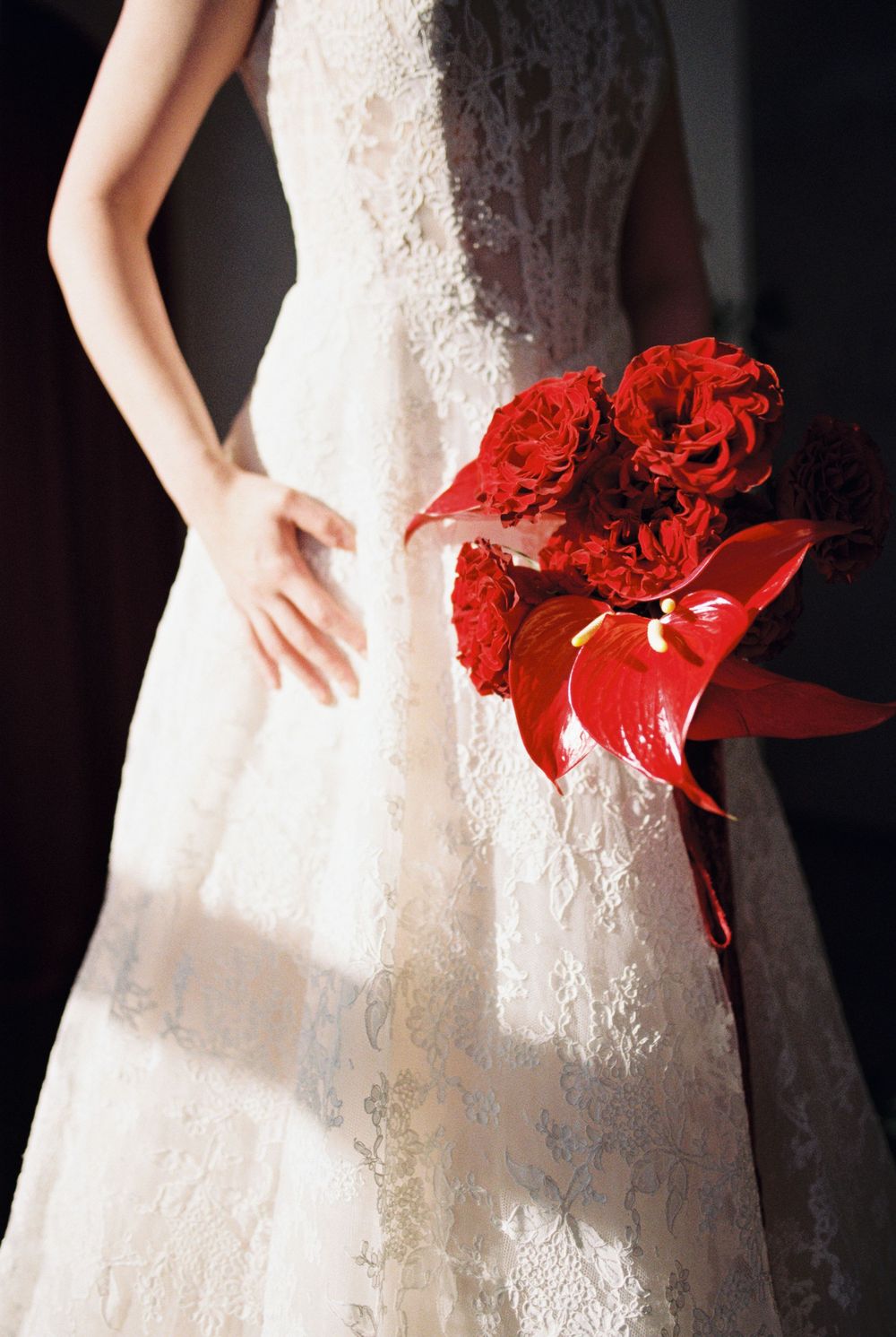 White lace wedding dress contrasts with vibrant red bouquet featuring roses and anthuriums in dramatic lighting.