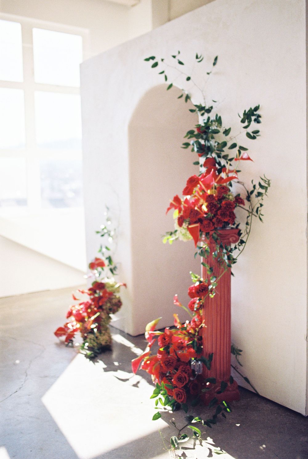 Bright red floral arrangements cascade down white arched walls in an ethereal sunlit room with deep shadows.