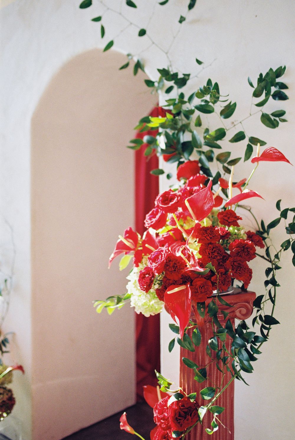 Elegant floral arrangement with red lilies and cascading greenery against a white wall with moody lighting.