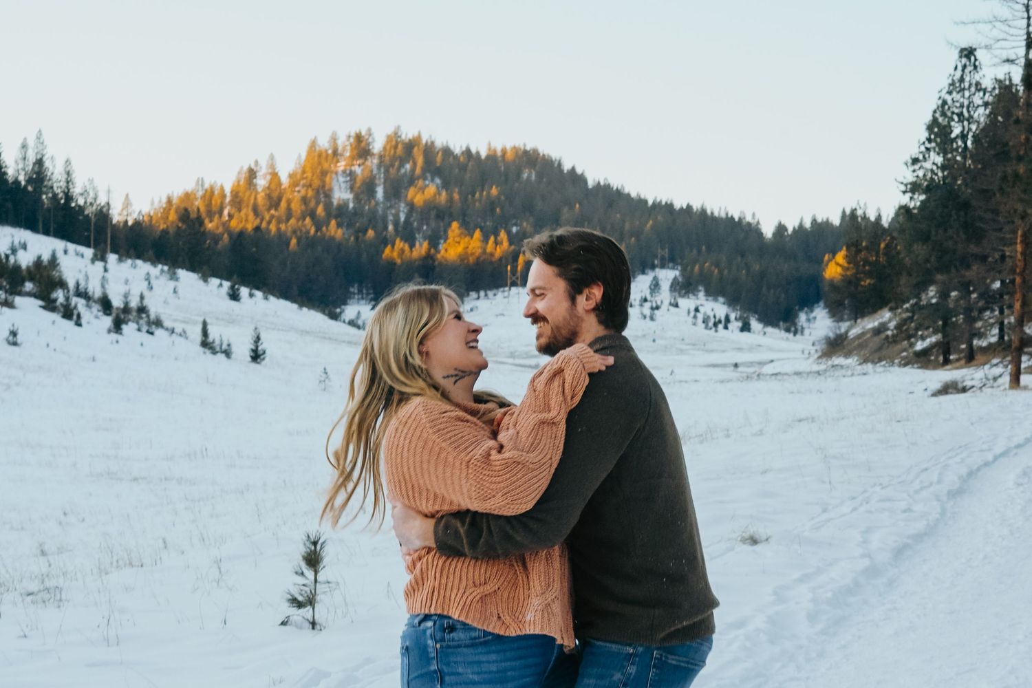 Romantic couple in warm winter clothing embrace in snowy mountain landscape at sunset.