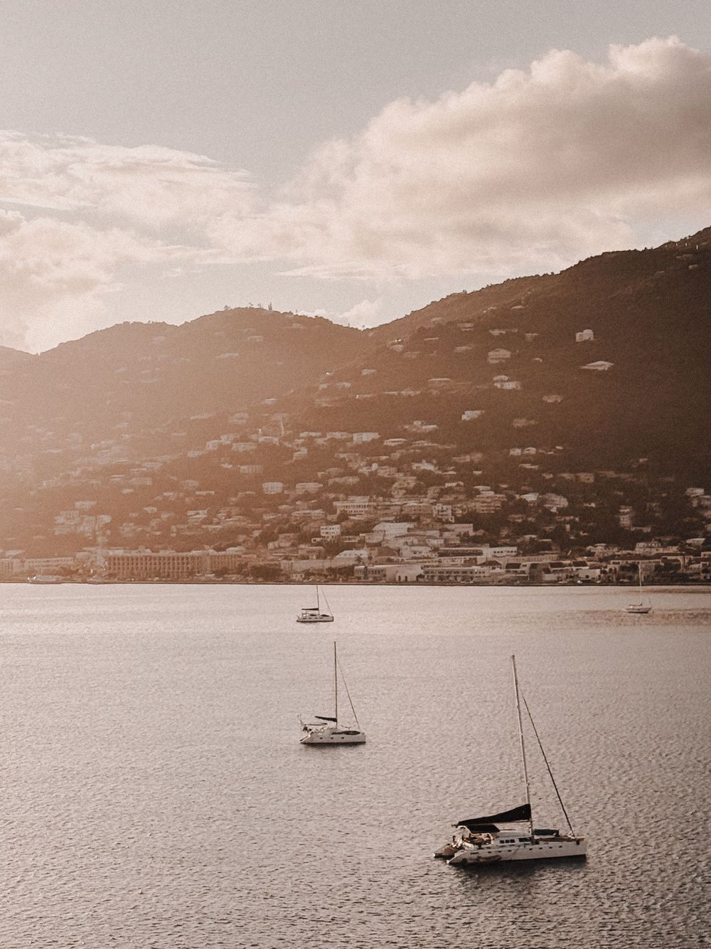 Coastal landscape with sailboats anchored in harbor against hillside homes at sunset.