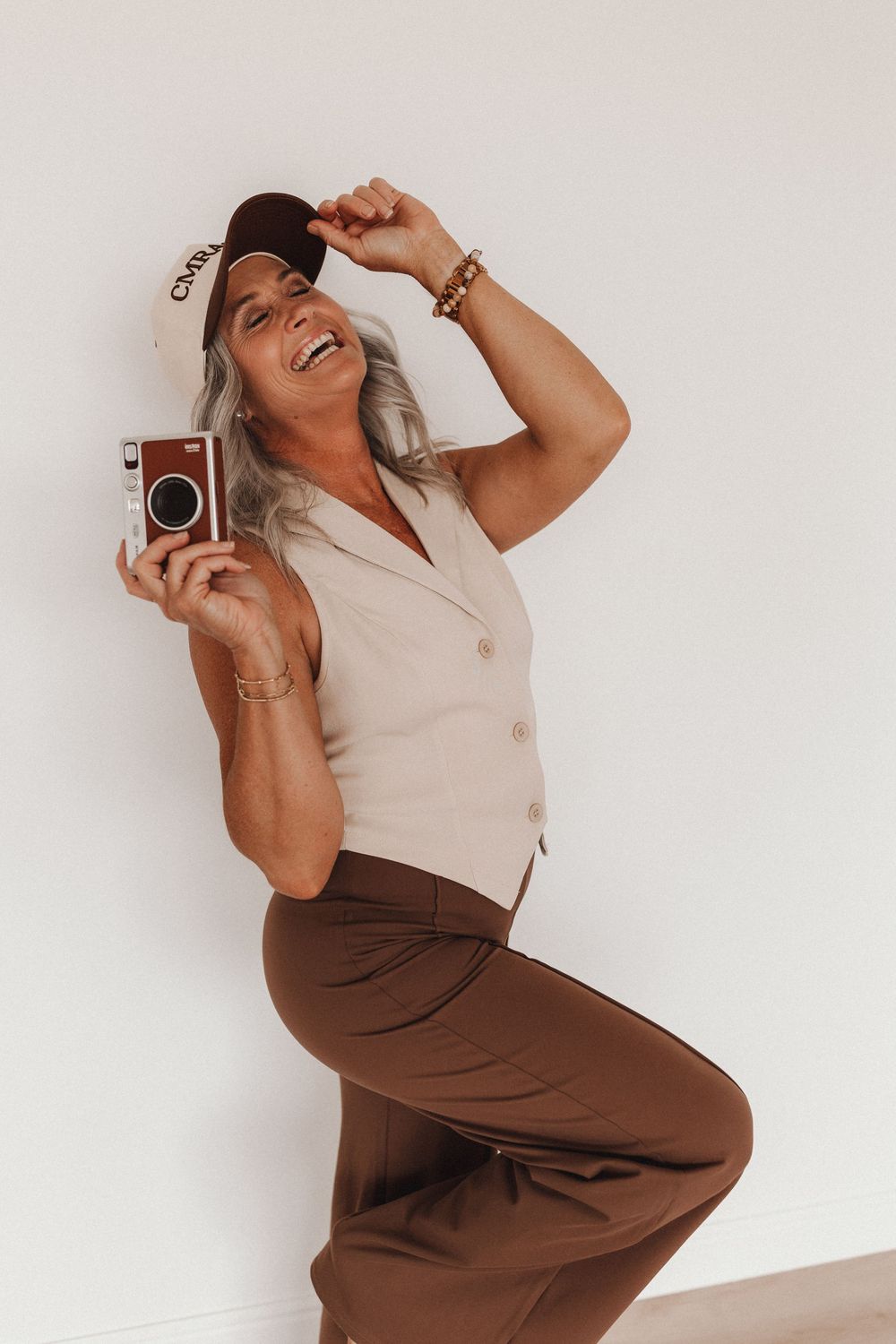 Retro-styled photographer in beige vest and brown pants poses playfully with vintage camera.