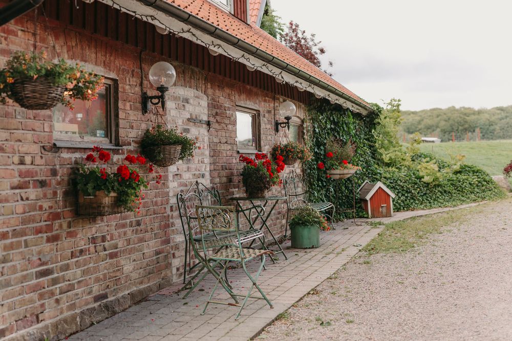 Rustic brick cottage exterior with hanging red flowers and metal bistro chairs along a gravel path in countryside setting.