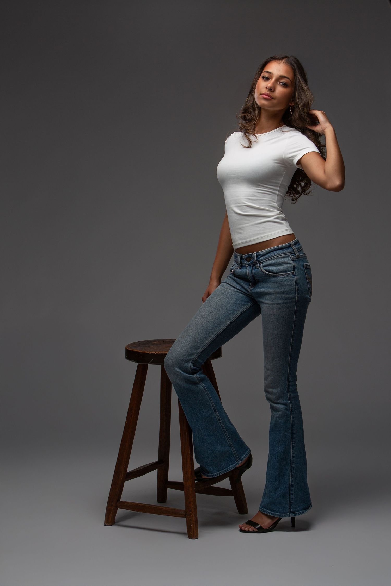 Fashion portrait series showing casual style with blue jeans and white top while posing on wooden stool against gray backdrop.