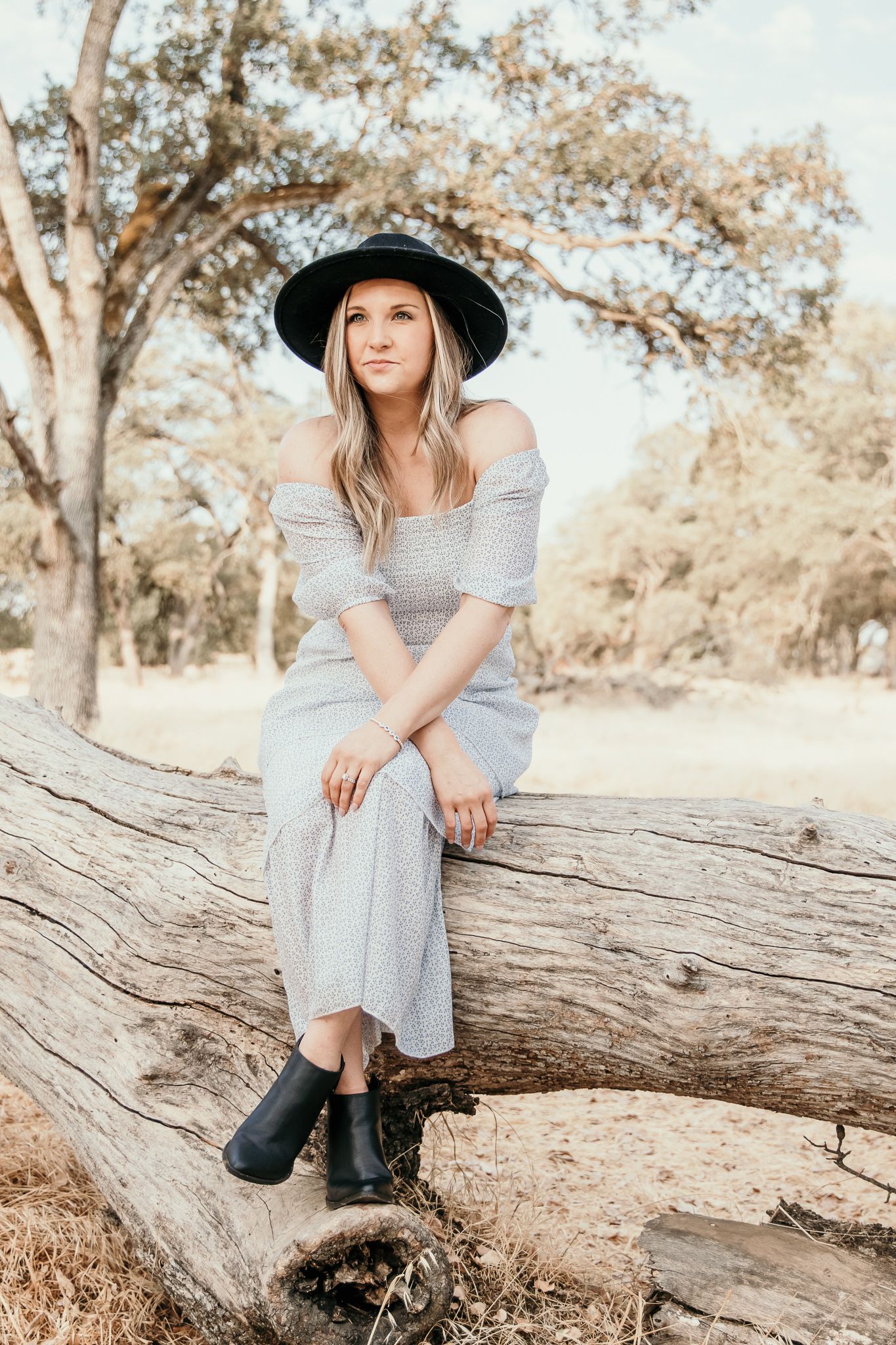 A person wearing a gray dress and black hat sits on a large fallen log in a sun-dappled outdoor setting.