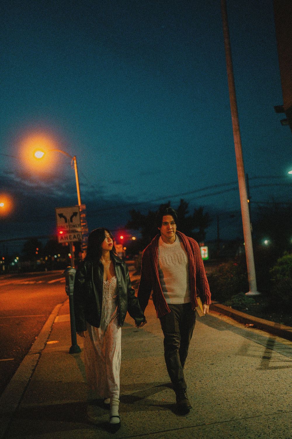 Two figures stroll along a dimly lit street path against a dusky blue sky with glowing streetlights.