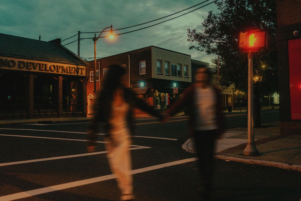 Blurred figures cross a moody urban street at dusk with neon lights and storefronts glowing in the background.