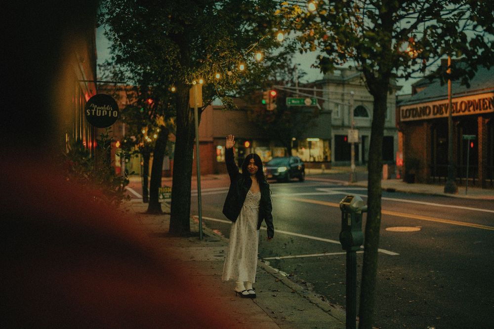 A moody urban street scene at dusk with warm street lights illuminating trees and buildings on a quiet corner.