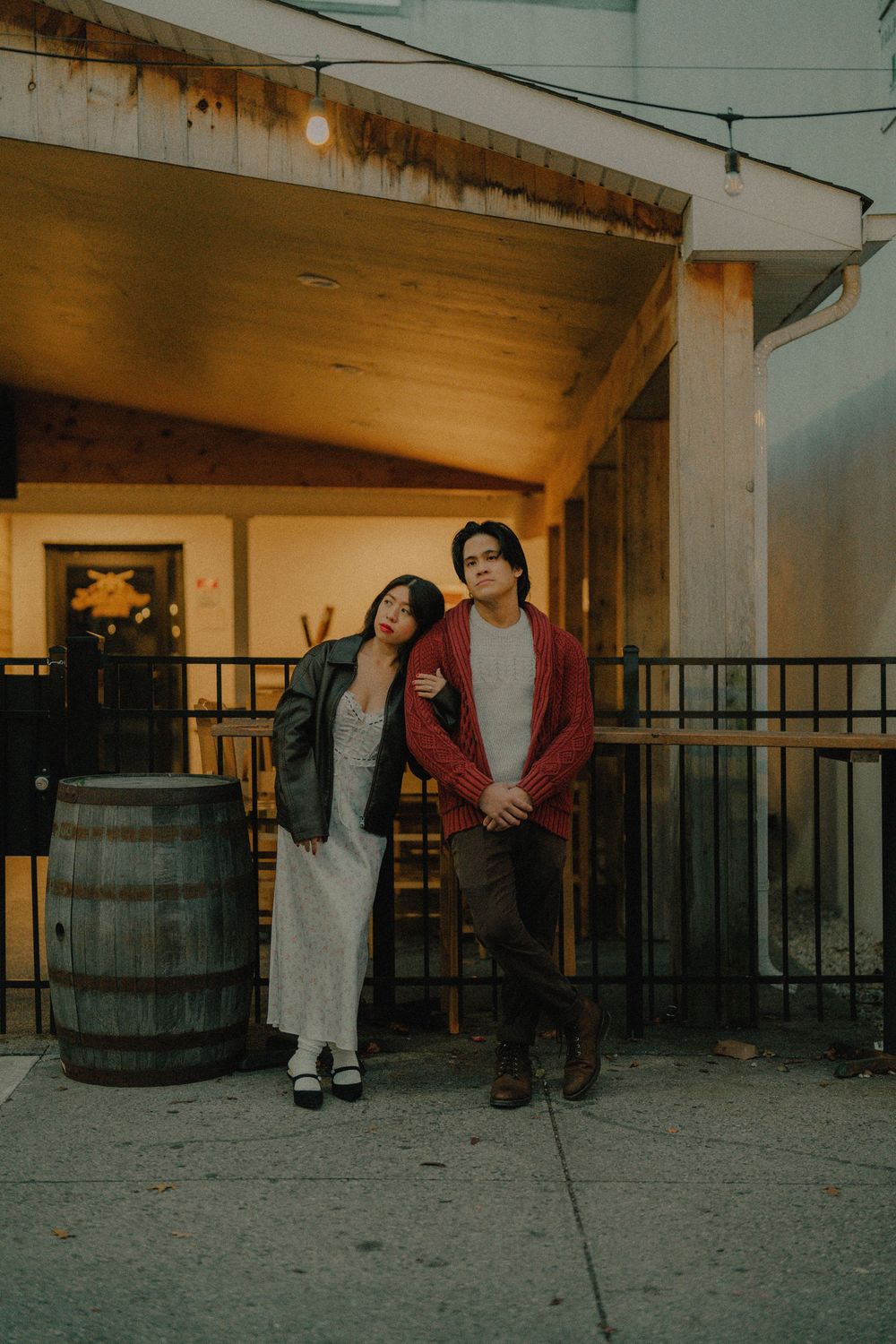 People walk past wooden barrels on a concrete porch at dusk.