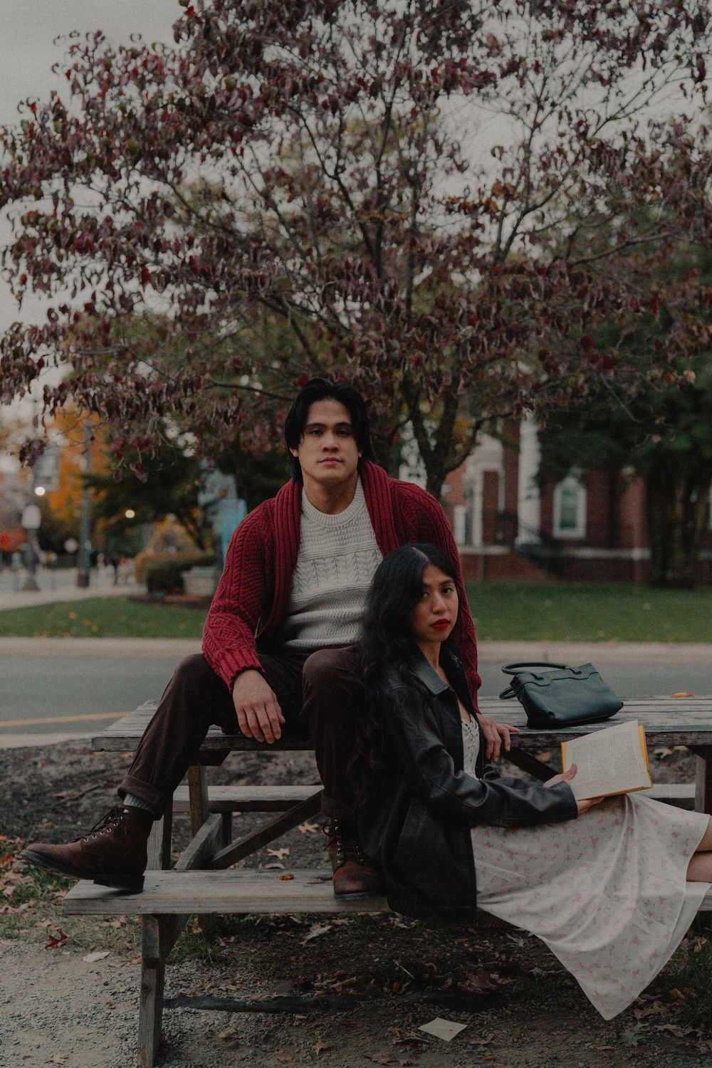 A couple sits together on a bench surrounded by autumn trees on a moody fall day.