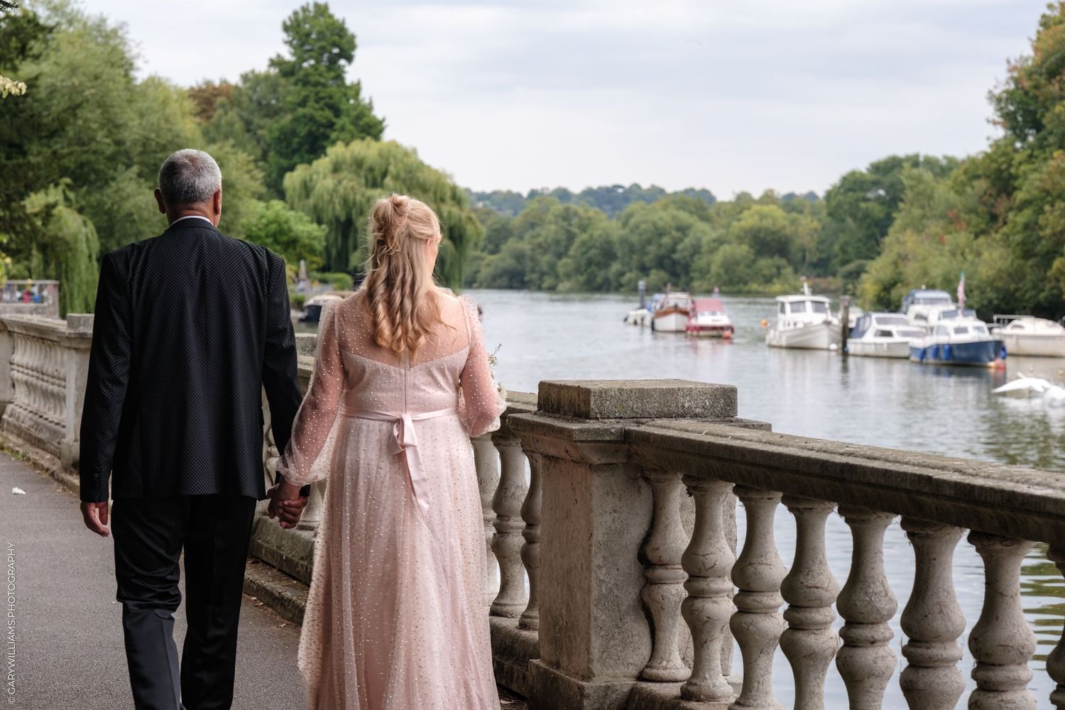 A couple stands at a stone balustrade overlooking a scenic river with moored boats and lush greenery.