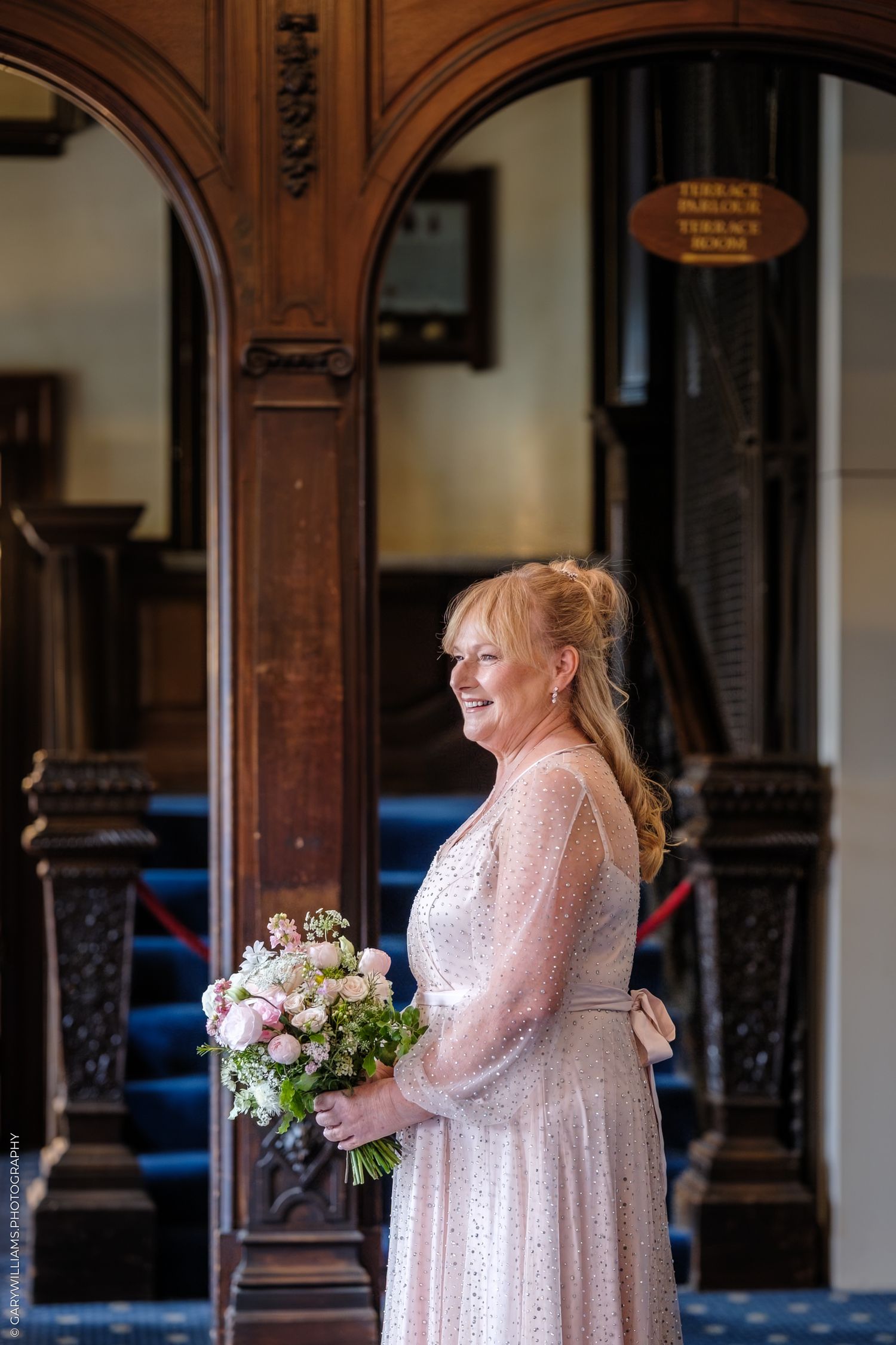 A person in a soft pink lace dress holds a delicate bouquet of flowers while standing in an ornate wooden hall.