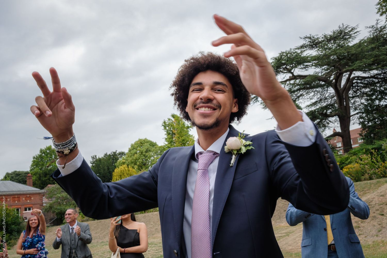 A cheerful wedding guest in a navy suit and purple tie makes celebratory hand gestures outdoors.