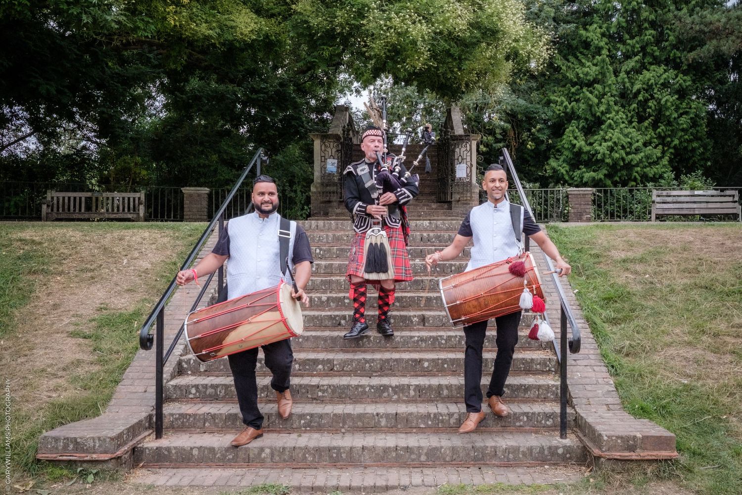 Musicians hold traditional drums while posing on stone steps in an outdoor park setting.