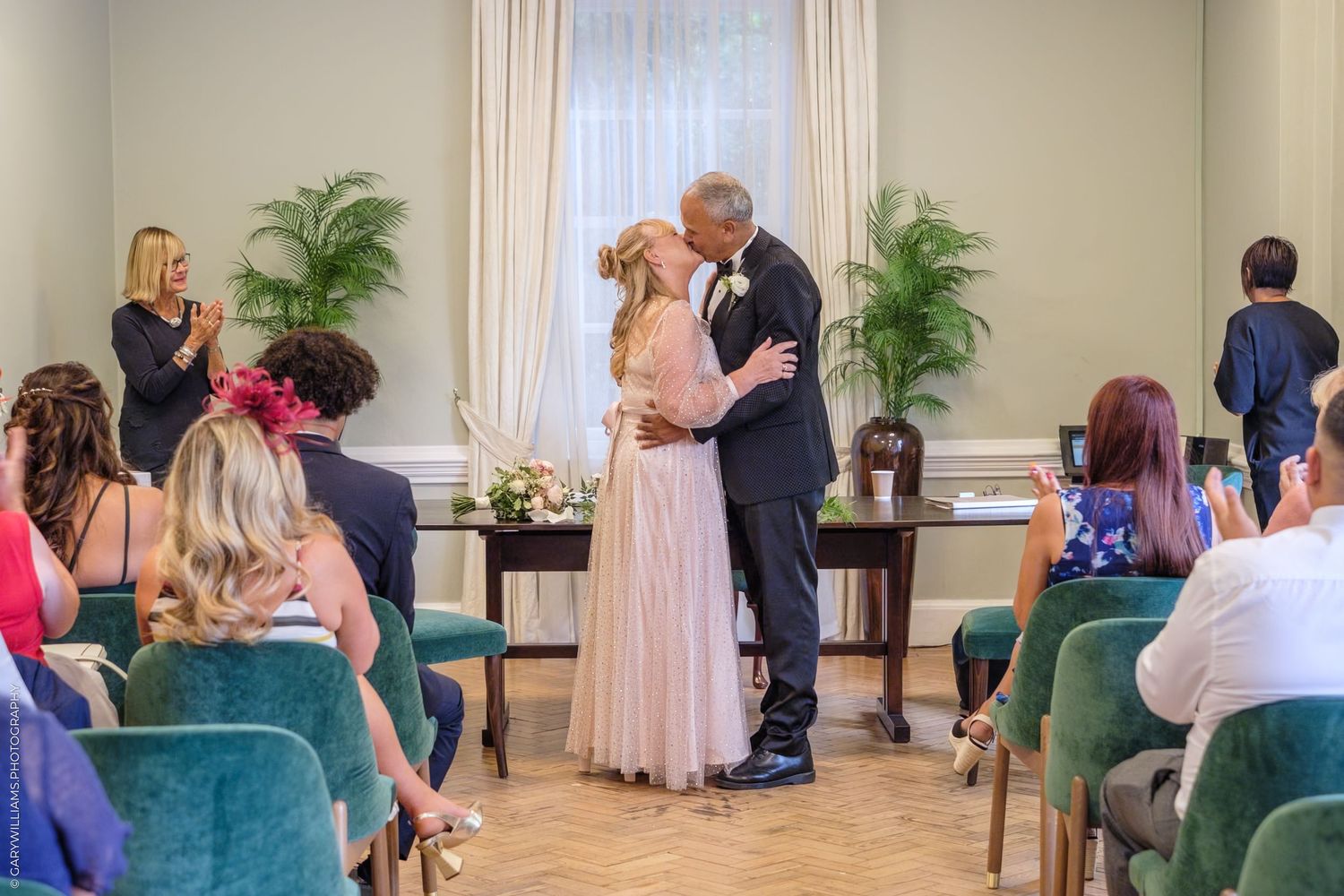 Wedding ceremony guests gather in a bright indoor venue with decorative plants and white curtains on the windows.