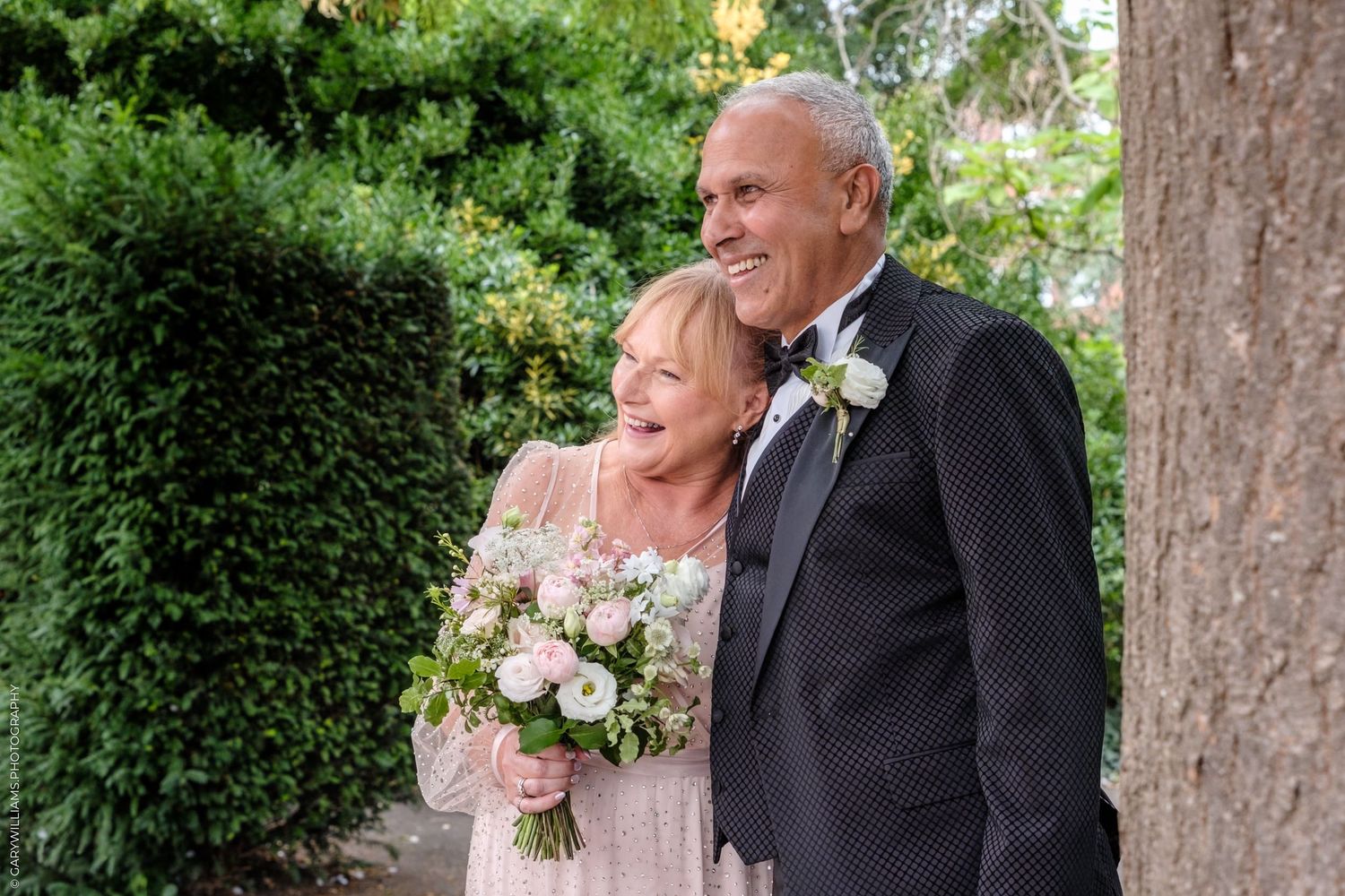 A wedding couple stands under tall trees outdoors holding pink bouquets during their summer garden ceremony.