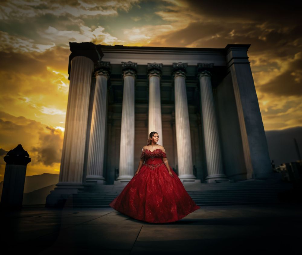 Dramatic sunset silhouettes a figure in a red ball gown standing before towering Greek-style columns of a classical building.