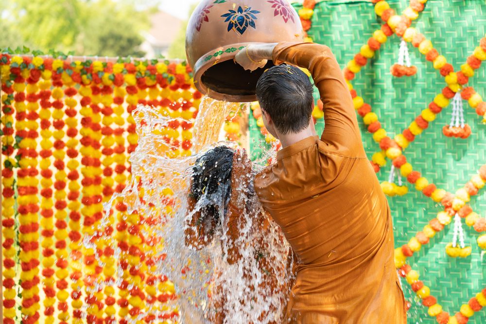 A person in an orange sweater splashes water on their face against a festive holiday light background. Haldi Quad Cities Photography