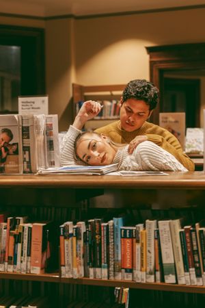 Students sleeping with their heads down on library desks surrounded by shelves of academic books.