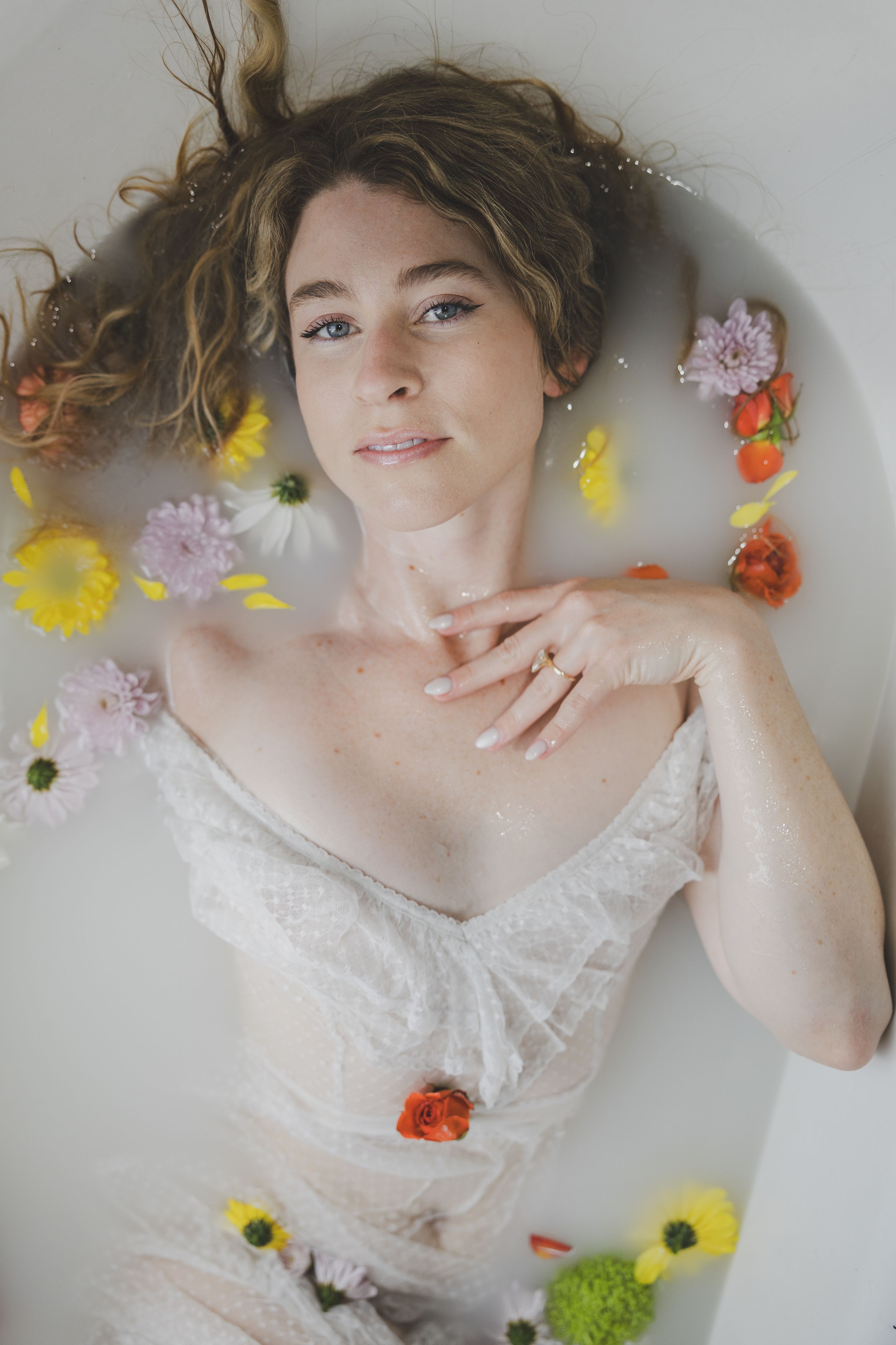 Ethereal photos feature person in white lace dress lying down surrounded by colorful scattered flowers and petals.