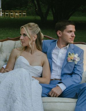 A couple in formal wedding attire sitting together on a stone wall during an outdoor photoshoot.