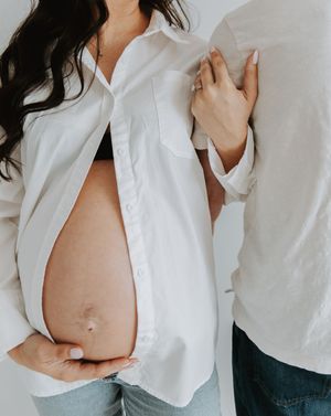 Close-up maternity photo showing a pregnant belly underneath an open white button-down shirt against a neutral background.