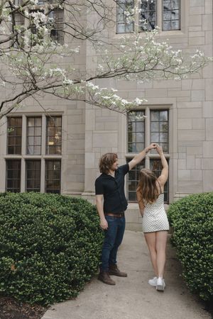 Couple dancing outdoors near a stone building with blooming white dogwood tree and trimmed hedges in spring.