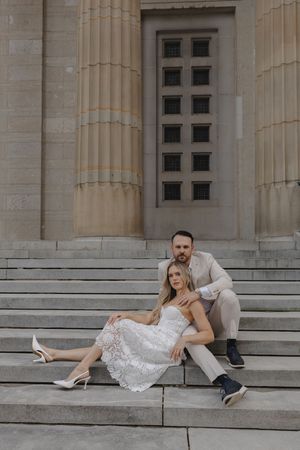 Bride and groom sitting on stone steps of a grand building, she in white lace dress, he in beige suit.