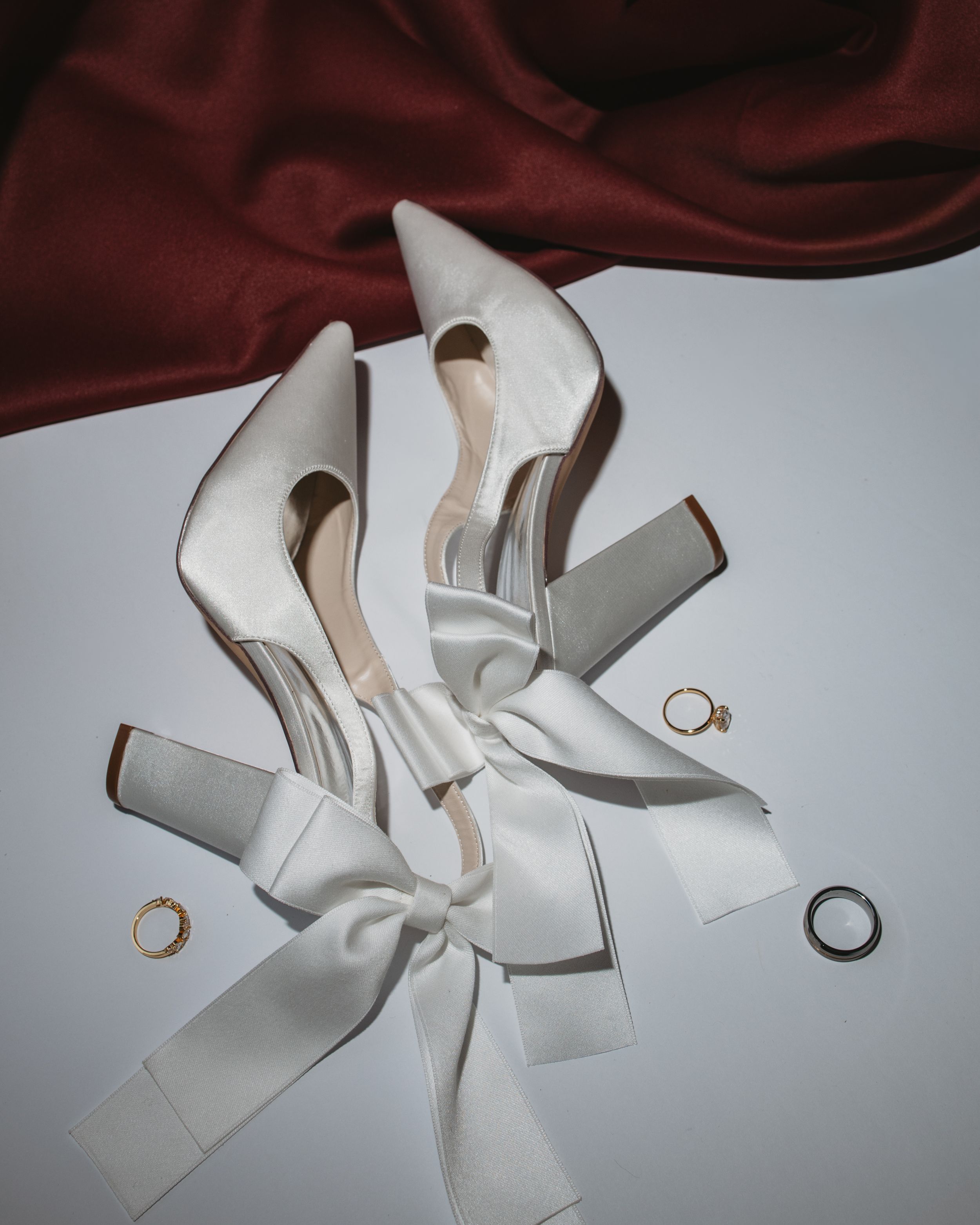Elegant white satin pointed-toe heels with bow ribbon, gold rings, and red silk fabric backdrop for a bridal flat lay.