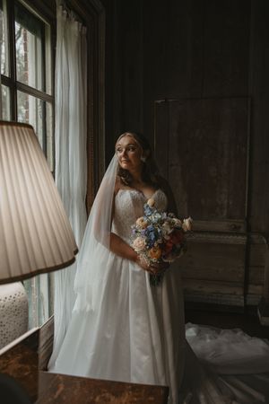 Bride in white gown and veil holds blue floral bouquet by window in elegant dark wood room.